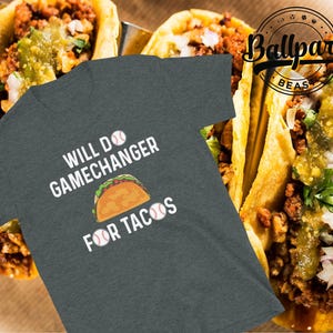 May include: Dark gray t-shirt with the text "Will Do Gamechanger For Tacos" and a taco graphic. Two baseballs are on either side of the word "tacos". The background features three tacos with salsa.