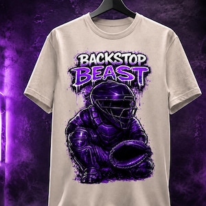 May include: A beige t-shirt featuring a purple graphic of a baseball catcher in full gear, with the words "BACKSTOP BEAST" in a stylized font above the illustration. The design has a distressed, paint-splatter effect.