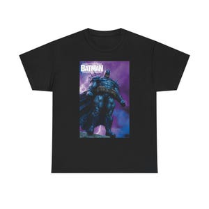 May include: Black t-shirt featuring a graphic of Batman in a blue and purple comic book style. The word "BATMAN" is printed in white at the top of the graphic. The superhero is depicted standing with a cape and spiked armor.