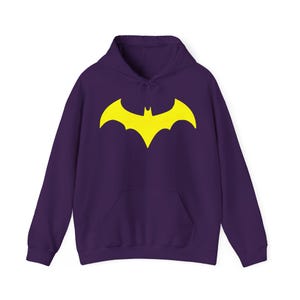 May include: A purple hoodie featuring a yellow Batman logo. The sweatshirt has a front pocket and a drawstring hood. The bat symbol is prominently displayed on the chest. The garment is made of a soft material.
