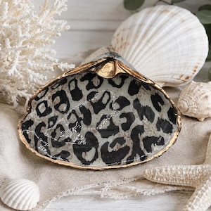 May include: A decorative clam shell dish featuring a black and white leopard print pattern on the interior surface. The edges of the shell are finished with a metallic gold trim, providing a distinct contrast to the animal print design.