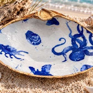 May include: A large, open seashell dish with a white interior featuring hand-painted blue illustrations of an octopus, a crab, and a snail. The edges of the shell are finished with a metallic gold trim, creating a decorative bowl for small items.