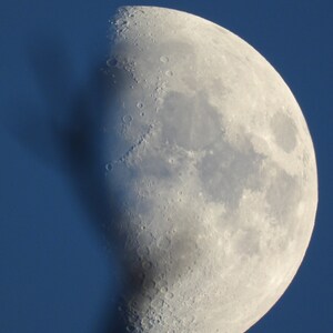 Könnte beinhalten: Eine Nahaufnahme eines zunehmenden Mondes vor blauem Himmel. Der Mond ist auf der rechten Seite beleuchtet und zeigt seine Krater und Oberflächenmerkmale.