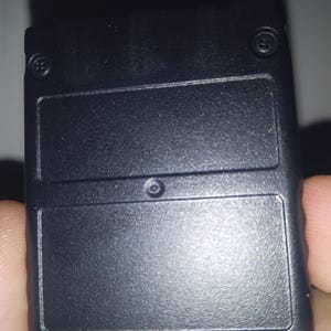 May include: A rectangular black plastic electronic game cartridge featuring a textured surface, two small screws at the top corners, and a central circular indentation. The bottom edge has a series of horizontal grooves molded into the casing.