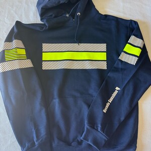 Puede incluir: Una sudadera con capucha azul marino con una franja reflectante amarilla en el pecho y una franja reflectante más pequeña en cada manga. Las mangas también presentan una bandera estadounidense blanca y el texto "Nevada Sunshine" en la manga derecha.