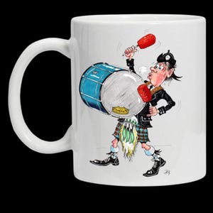 May include: A white ceramic mug featuring a colourful cartoon illustration of a drummer wearing a kilt and a black hat. The character is playing a large blue bass drum with red mallets, showing a dynamic pose with visible sweat and motion lines.