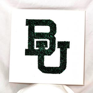 May include: Square white canvas featuring the letters "BU" in dark green rhinestones. The letters are large and centered, with the rhinestones creating a sparkling effect. The background is plain white.