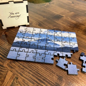Pode incluir: Um quebra-cabeça de madeira de uma cena de lago de montanha com algumas peças faltando. O quebra-cabeça está sobre uma mesa de madeira ao lado de uma pequena caixa de madeira com as palavras "You are Amazing!" gravadas nela.