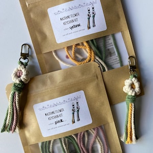 May include: Two macrame flower keychain kits in brown paper packaging. One kit is labeled "yellow" and the other "pink". Each kit includes materials and instructions. Finished keychains with flower designs are also visible.