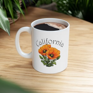 May include: White ceramic coffee mug with a black handle. The mug has a graphic of two orange California poppies with green stems and leaves. The text "California" is printed in black on the mug.