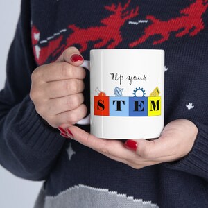 May include: White ceramic mug with a blue, red, yellow, and green design. The mug says "Up your STEM" with a beaker, satellite, gear, and calculator graphic.