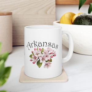 May include: White ceramic coffee mug with a black and white image of pink flowers and the word "Arkansas" printed on the front.