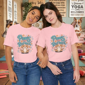 May include: Two people wearing pink t-shirts with a graphic of a dog meditating with the text "Paws and Reflect" in blue and green lettering. The shirts are decorated with flowers and leaves.