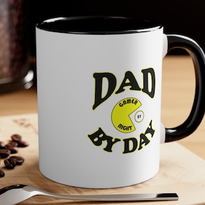 May include: White ceramic mug with a black rim and handle. The mug features a black and yellow graphic that says "Dad Gamer by Night By Day".
