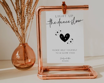 May include: A white sign with black text and a black heart with stars inside. The sign says "Light up the dance floor" and "Please help yourself to a glow stick." It also includes the names "Emily and Alex" and the date "26.11.20XX". The sign is displayed on a copper stand.