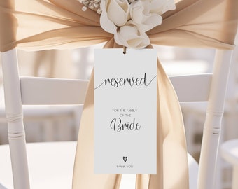 May include: White card with black text that says "reserved" for the family of the bride. The card is tied to a white chair with a beige sash and white flowers.