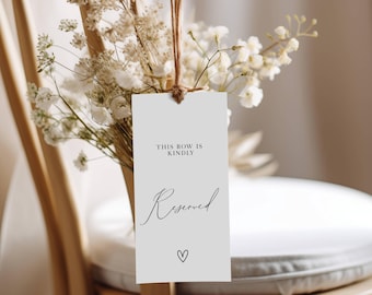 May include: White card with the text "This row is kindly reserved" and a small heart symbol hanging from a wooden chair with dried white flowers.