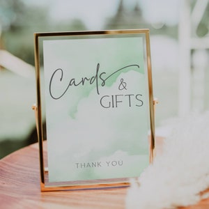 May include: A gold-framed sign with a green watercolour background. The sign reads "Cards & Gifts" with the text "Thank You" at the bottom.