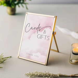 May include: A gold-framed sign with a pink watercolor background. The sign reads "Cards & Gifts" with the text "Thank You" at the bottom.