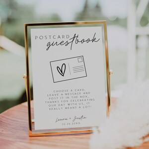 May include: A white sign with black text and a black and white illustration of a postcard. The sign reads "Postcard Guestbook" and includes instructions for guests to leave a message on a postcard. The sign also includes the names "Laura + Justin" and the date "10.26.20XX".