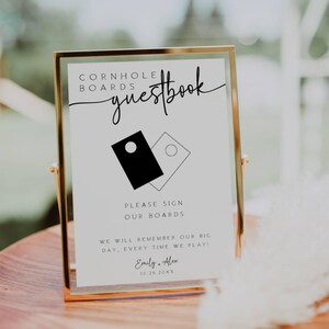 May include: A white sign with black text that reads "Cornhole Boards Guestbook". The sign features a black and white illustration of two cornhole boards. The text below the illustration reads "Please sign our boards. We will remember our big day, every time we play! Emily & Alex 10.26.20XX".