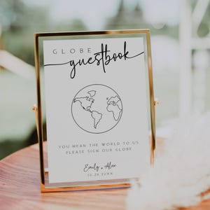 Pode incluir: Uma placa com moldura dourada com o texto "GLOBE guestbook" e um desenho de linha do mundo. O texto abaixo diz "You mean the world to us, please sign our globe." Os nomes "Emily + Alex" e a data "10.26.20XX" estão impressos abaixo.