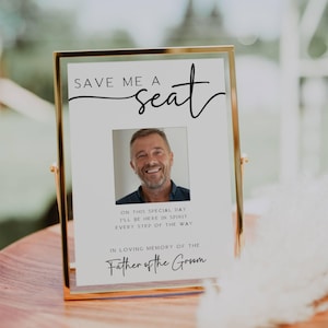 以下が含まれることがあります： 「Save Me A Seat」と黒文字で書かれた白い看板に、金色の額縁に入った男性の写真が飾られています。写真の下には、「この特別な日に、私は心の中であなたと一緒です。すべての歩みを共にします。新郎の父の思い出に。」と書いてあります。