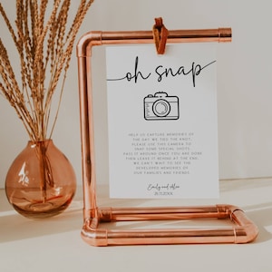 May include: White card with the text "oh snap" in black cursive font. A black and white camera icon is below the text. The card also includes the text "Help us capture memories of the day we tied the knot, please use this camera to snap some special shots. Pass it around once you are done then leave it behind at the end. We can't wait to see the developed memories of our families and friends. Emily and Alex 26.11.20XX". The card is displayed on a copper pipe stand.
