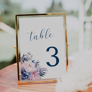 May include: A gold-framed table number sign. The sign displays the word "table" in navy script and the number "3" in a bold navy font. It features a floral design with pink and purple flowers.