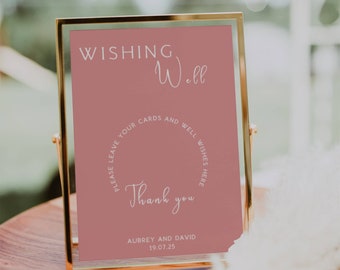 May include: A framed sign with the words "Wishing Well" in white script. The sign is a dusty rose color with a circular text that reads "Please leave your cards and well wishes here." The sign also says "Thank you" and the names "Aubrey and David" with the date "19.07.25".