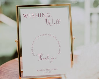 May include: A gold-framed sign with the text "WISHING Well" in pink script. The sign also includes the text "PLEASE LEAVE YOUR CARDS AND WELL WISHES HERE" in a circle around the words "Thank you". The bottom of the sign reads "AUBREY AND DAVID 19.07.25".