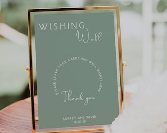 May include: A gold-framed sign with a sage green background and white text that reads "WISHING Well PLEASE LEAVE YOUR CARDS AND WELL WISHES HERE Thank you AUBREY AND DAVID 19.07.25".