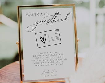 May include: A white sign with a gold frame, with the text "POSTCARD guestbook" in black script. The sign has a black and white illustration of a postcard with a heart on it. The text below the illustration reads "CHOOSE A CARD, LEAVE A MESSAGE AND POST IT IN THE BOX. THANKS FOR CELEBRATING OUR DAY WITH US, IT REALLY MEANS A LOT!" with the names "Balding & Jane" and the date "10.26.20XX" below.