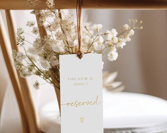 May include: White card with gold text that reads "This row is kindly reserved" with a small gold heart at the bottom. The card is hanging from a wooden chair with dried white flowers.