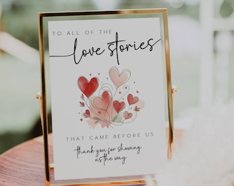 May include: A white sign with a gold frame, featuring a watercolor illustration of red hearts and the text "TO ALL OF THE love stories THAT CAME BEFORE US thank you for showing us the way".