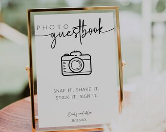 May include: A gold-framed sign with a white background and black text that reads "PHOTO guestbook". A black and white illustration of a camera is above the text. The text below the camera reads "SNAP IT, SHAKE IT, STICK IT, SIGN IT." The names "Emily and Alex" and the date "26.11.20XX" are at the bottom of the sign.