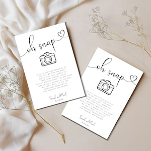 Puede incluir: Dos tarjetas blancas con texto negro y un icono de cámara simple. El texto en las tarjetas dice "oh snap" y pide a los invitados que usen la cámara para tomar fotos en la boda. Las tarjetas están firmadas por "Sarah y Mark" e incluyen la fecha "26.11.20XX".