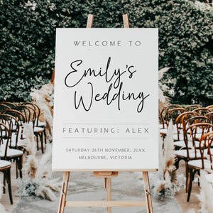 Może przedstawiać: Biały znak powitalny na weselu na drewnianym sztaludze. Znak głosi: "WELCOME TO Emily's Wedding FEATURING: ALEX SATURDAY 26TH NOVEMBER, 20XX MELBOURNE, VICTORIA." W tle widać rzędy krzeseł i zieleń.