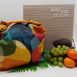 Könnte beinhalten: Eine wiederverwendbare Lunch-Tasche mit einem farbenfrohen geometrischen Muster in Blautönen, Grün, Gelb, Orange und Rot. Die Tasche ist aus Stoff und hat einen Druckknopfverschluss. Die Tasche liegt auf einer weißen Oberfläche mit einem Holzschild, auf dem "Mona Kind Designs" in weißen Buchstaben steht.
