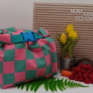 Könnte beinhalten: Ein wiederverwendbarer Lebensmittelverpackung aus rosa und grünem Karomuster mit einer blauen Schnalle. Die Verpackung ist gefaltet und mit einem grünen Band gebunden. Die Verpackung liegt auf einer weißen Oberfläche mit einem Farn, Karotten, Himbeeren und einer Avocado.