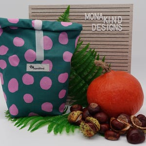 Könnte beinhalten: Eine wiederverwendbare Lunch-Tasche in Tealblau mit rosafarbenem Punkte-Muster. Die Tasche hat ein weißes Etikett mit dem Text "monakind" darauf. Die Tasche liegt auf einer weißen Oberfläche mit einem Kürbis, Kastanien und grünen Farnen.