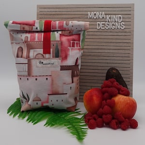 Könnte beinhalten: Eine wiederverwendbare Lunchtasche mit einem farbenfrohen Druck einer Stadtlandschaft mit roten, weißen und braunen Gebäuden. Die Tasche hat einen roten Riemen und der Markenname "monakind designs" ist sichtbar. Frisches Obst, darunter Äpfel, Himbeeren und eine Avocado, sind in der Nähe angeordnet.