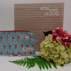 Könnte beinhalten: Eine blaue Reißverschlusstasche mit einem Muster von Schwimmern in roten Badeanzügen. Die Tasche hat einen roten Reißverschluss und ein kleines Etikett mit der Aufschrift "mona kind". Ein Holzschild mit der Aufschrift "MONA KIND DESIGNS".
