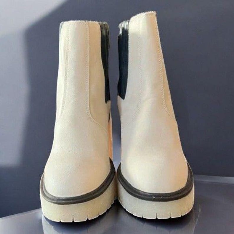 May include: A pair of white ankle boots with black trim around the top of the boot and a black elastic panel on the back of the boot.
