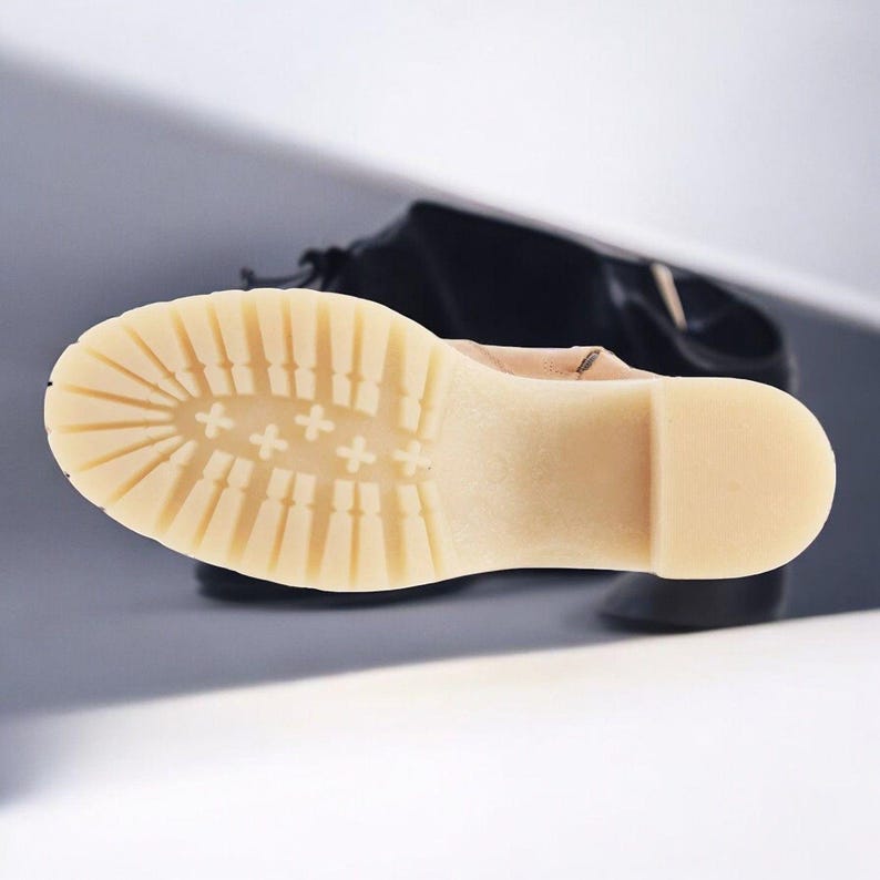 May include: Close-up of the bottom of a light brown leather boot with a cream-coloured sole. The sole has a tread pattern with multiple rows of raised bumps and a series of plus signs.