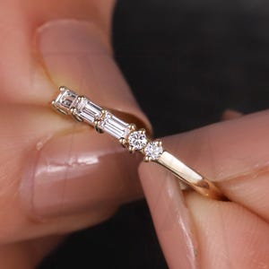 Puede incluir: Un delicado anillo de oro con una serie de pequeños diamantes brillantes. El anillo tiene una banda delgada y está adornado con una combinación de diamantes de talla baguette y redondos, creando un diseño refinado y elegante. El anillo se sostiene entre dos manos.