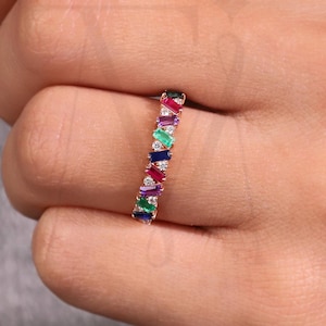 以下が含まれることがあります： 赤、青、緑、紫の長方形の宝石が並んだゴールドリング。 リングは手の上にあり、灰色の背景に設定されています。