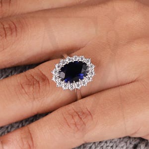 Könnte beinhalten: Ein silberner Ring mit einem großen ovalen, dunkelblauen Edelstein, umgeben von einem Kranz aus kleineren, klaren Steinen. Der Ring befindet sich an einer Hand, im Hintergrund ein grauer Pullover. Der Ring ist ein feines Schmuckstück.