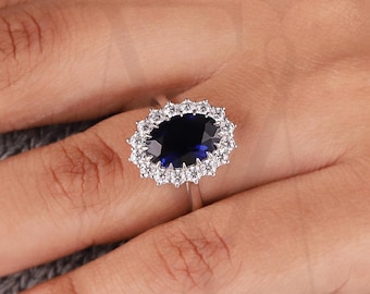Könnte beinhalten: Ein silberner Ring mit einem großen ovalen, dunkelblauen Edelstein, umgeben von einem Kranz aus kleineren, klaren Steinen. Der Ring befindet sich an einer Hand, im Hintergrund ein grauer Pullover. Der Ring ist ein feines Schmuckstück.