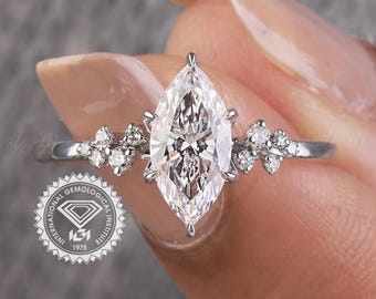 Op de afbeelding: Zilveren verlovingsring met een marquise-geslepen centrale steen, geflankeerd door kleinere stenen. De ring wordt door een hand omhoog gehouden. Een luxe sieraad, ideaal voor een huwelijksaanzoek.